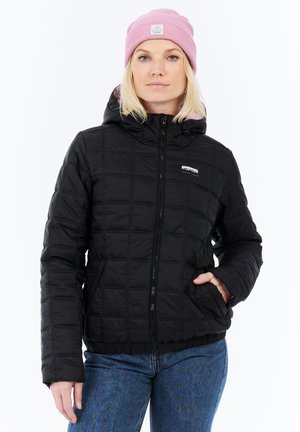 Vrouw in een zwarte gewatteerde jas met capuchon, blauwe jeans en een roze beanie, staande met een hand in de zak van haar jas tegen een witte achtergrond.