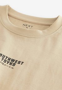 Beige T-paita, jossa on pyöreä kaula-aukko, mustalla printatulla logolla "SOUTHWEST TOYKO" ja lisäteksti. Pehmeä, tekstuuri puuvillamateriaali.