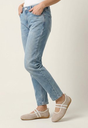 Person iført lyseblå jeans og beige flade sko med hvide remme, stående med hænderne i lommerne foran en ensfarvet baggrund.