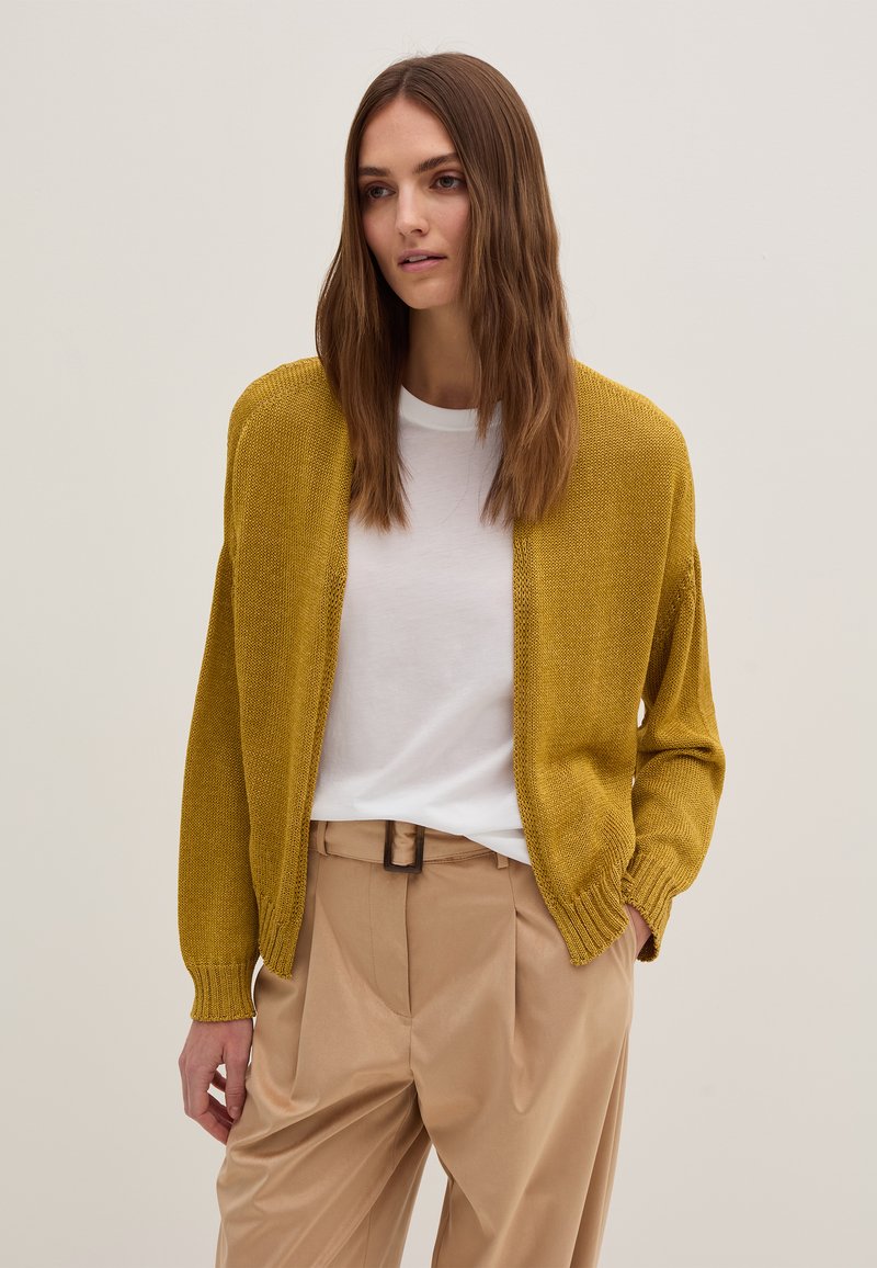 Cárdigan de punto color mostaza con un corte suelto, puños acanalados, combinado con una camiseta blanca y pantalones plisados de color crema.