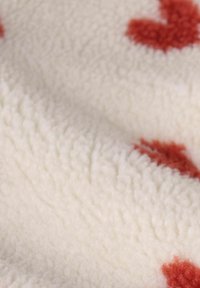 Tissu en polaire blanche présentant une texture douce avec des motifs de cœurs rouges. Le matériau a une sensation épaisse et pelucheuse avec des détails de fibres visibles.