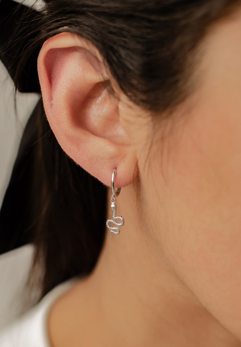 Pendientes de aro de plata con un charm de serpiente con un detallado intrincado, colgando junto al lóbulo de la oreja sobre un fondo difuminado.