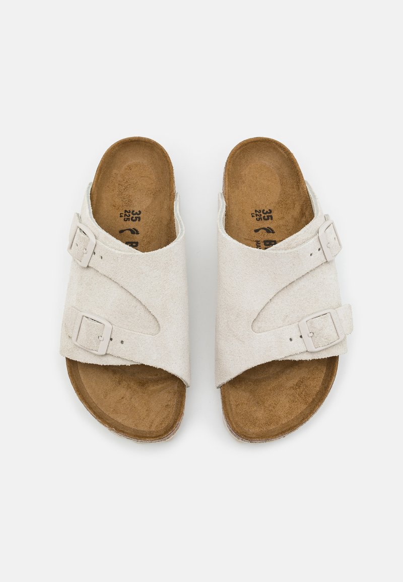 Helle beige Wildledersandalen mit zwei verstellbaren Riemen und metallischen Schnallen. Die konturierte braune Innensohle bietet Unterstützung. Flaches, offenes Design mit offenem Zehenbereich.