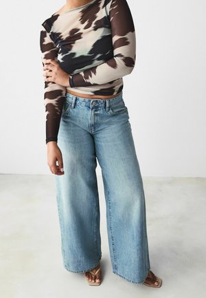 Langärmlige, durchsichtige Bluse mit einem braun-creamfarbenen abstrakten Muster, kombiniert mit hoch taillierten, weit geschnittenen hellblauen Jeans und nude Farbene Sandalen.
