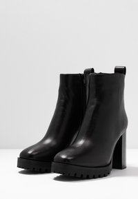 Mustat nahkakengät nilkkoihin, joissa on sileä pinta, pyöreä kärki, sivuvetoketju ja paksu, teksturoitu pohja. Varusteena korkea, lohkareen muotoinen korokepohja.