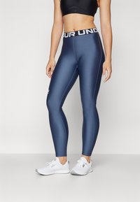 Námornícke modré legíny s hladkou, elastickou textúrou, s čiernym pásom a logom "UNDER ARMOUR". Skombinované s bielymi športovými topánkami.