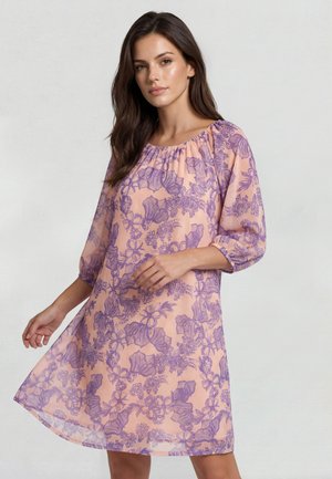 Ung kvinde iført en løs ferskenfarvet kjole med lilla blomsterprint og trekvartærmer, stående foran en ensfarvet lys baggrund.