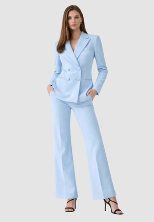 Donna che indossa un blazer doppiopetto azzurro chiaro e pantaloni larghi abbinati con sandali neri con tacco alto, in piedi con le mani in tasca.
