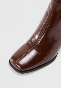 Bota de tornozelo em couro sintético castanho com bico pontudo, textura lisa e detalhes de costura visíveis ao longo do lado. Sola preta.