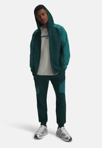 Grüner Kapuzen-Sweatshirt mit Reißverschluss, graues T-Shirt darunter, passende Hose mit Paneeldetails und silberne Sportschuhe.