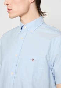 Camisa azul-clara de botão, feita de tecido de algodão, com um bolso no peito apresentando um logótipo bordado e um design de colarinho clássico.
