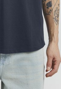 Marineblå bomull t-skjorte med rund hals, korte ermer og rett nederkant, sammen med lysvaskede denim jeans. Synlige tattooer på armen.