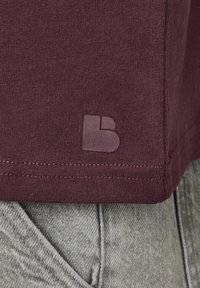 T-shirt bordeaux avec une texture lisse et un col rond doux, comportant une étiquette en tissu gris et des coutures contrastantes à l'ourlet.