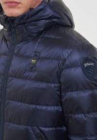 Giacca puffa blu navy con cappuccio, caratterizzata da un design trapuntato, tasche con zip e toppe con logo sul petto e sul braccio. Texture liscia.