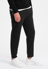 Schwarze Sweatpants mit schlanker Passform, ausgestattet mit Nahtdetails und seitlichen Reißverschlüssen, kombiniert mit weißen Sneakers. Glatte Textur und elastische Bündchen.