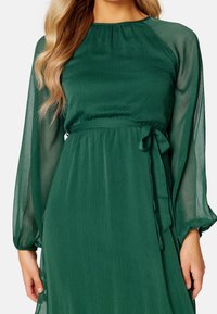 Vestido verde oscuro con una textura satinada, mangas largas transparentes, escote fruncido y un detalle de lazada en la cintura para ajustar.