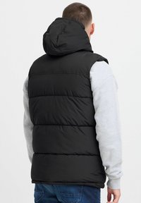Schwarze Pufferweste mit Kapuze, mit horizontalem Steppdesign und Reißverschluss. Trägt man über einem grauen Langarmshirt.