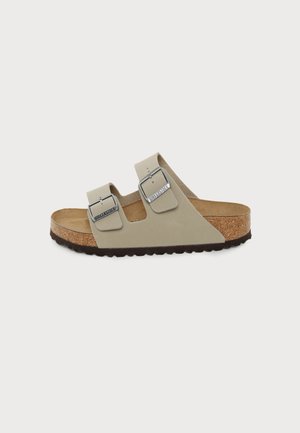 Béžové Birkenstock sandále s dvomi remienkami, nastaviteľnými kovovými prackami, korkovou stielkou a čiernou textúrovanou podrážkou na bielom pozadí.