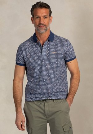 Man met kort haar en baard, gekleed in een blauw poloshirt met patroon en olijfgroene cargobroek, staande tegen een neutrale achtergrond.