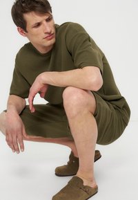 Άνδρας σκύβει φορώντας ελαιόχρωμο υφασμάτινο κοντομάνικο πουκάμισο και σορτς με καφέ σανδάλια χωρίς λουριά σε λευκό φόντο.