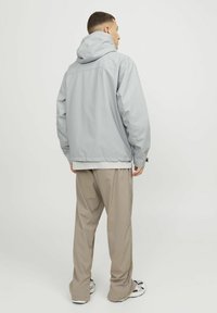 Hellgraue leichte Jacke mit Kapuze, die ein dezentes Karomuster aufweist, kombiniert mit lockeren beigen Hosen und sportlichen Schuhen.
