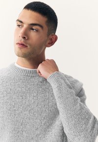 Maglione grigio lavorato a maglia con un motivo intrecciato e testurizzato. Presenta una scollatura rotonda e polsini a coste, realizzato con un materiale morbido e caldo.