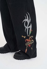 Svarta vida jeans med ett silvrigt flamdesign och en färgglad hjärt-tatuering med "PARIS" på nederdelen av benet; svarta skor med tjock sula.