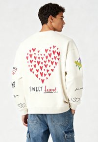 Krém színű pulóver nagy piros szívvel a hátsó részen, rajta a "SWEET heart" felirattal. A ujjakon különböző illusztrált díszítések találhatók.