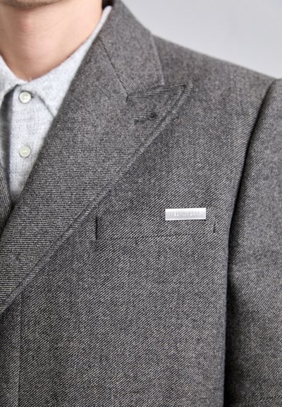 Hall herringbone bleiser hallide narmaga, rinna tasku ja hõbedane logo nõel. All pool on heledast hallist kraega särk. Tekstuuriga kangas.