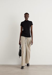 Zwarte polo, beige wijde broek, zwarte handtas en schoenen met een puntige neus. Model staat tegen een effen muur.