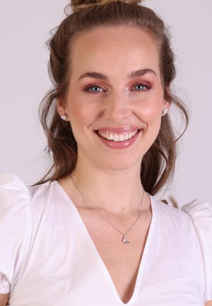 Silberne Halskette mit einem kleinen Anhänger; das Modell trägt eine weiße Bluse mit Puffärmel. Der Hintergrund ist hellgrau. Klare Haut und dezentes Make-up.