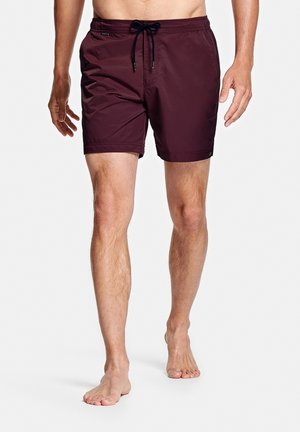 Man die blootsvoets loopt en donkere bordeauxrode zwemshorts met een zwarte koordsluiting draagt, op een witte achtergrond.