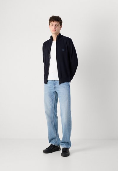 Chaqueta navy con cremallera y cuello alto, llevada sobre una camiseta blanca, combinada con jeans de mezclilla azul claro y zapatillas negras. Tejido suave, ajuste casual.