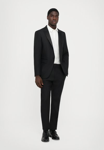 Schwarzer maßgeschneiderter Anzug mit einreihiger Jacke und flachen Hosen. Weißes Hemd. Glänzende schwarze Dressschuhe. Einfaches, klares Design.