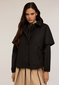 Jachetă neagră scurtă cu guler înalt, închidere cu nasturi și manșete elastice. Prezintă un design elegant și detalii cu buzunare cu fermoar.
