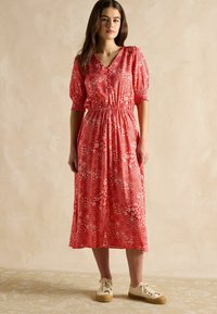 Rotes, blumenverziertes Midikleid mit puffärmel, geraffter Taille und V-Ausschnitt. Kombiniert mit cremefarbenen Sneakern vor einem neutralen Hintergrund.