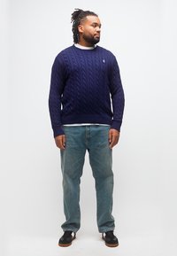 Άνδρας με πλεξούδες, φορώντας μπλε πουλόβερ με πλέξη καλώδιο, ανοιχτόχρωμο τζιν και μαύρα αθλητικά παπούτσια, στέκεται μπροστά σε λευκό φόντο, κοιτάζοντας προς τα δεξιά.