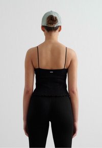 Svart ribbad linne med tunna band, scallopad nederkant, bärs med svarta leggings; person med en ljusblå keps vänd bort från kameran.