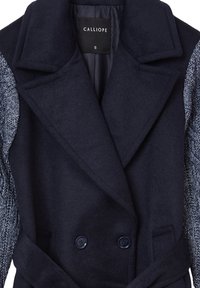 Cappotto in misto lana di colore blu navy con chiusura doppiopetto, grande colletto e maniche in maglia grigia testurizzata. Presenta bottoni e una cintura da annodare.