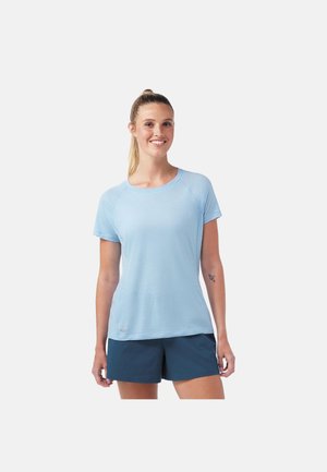 Jeune femme avec une queue de cheval blonde portant un t-shirt bleu clair à manches courtes et un short bleu foncé, souriante, debout devant un fond uni.