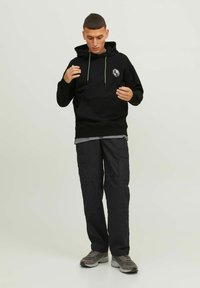 Un sweat à capuche noir avec un logo sur le devant, des cordons de serrage verts, superposé sur une chemise grise, accompagné d'un pantalon cargo noir et de baskets grises.