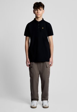 Teenager-Junge, der nach vorne steht, trägt ein schwarzes Polohemd, graue Cargohosen und weiße Turnschuhe vor einem schlichten grauen Hintergrund.