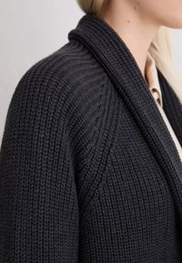 Dunkelgrauer Strickcardigan mit Schalkragen, geripptem Muster und offener Vorderseite, die die aufwendigen Nähte betont.