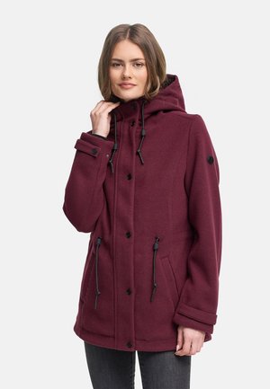 Világosbarna hajú nő bordó kapucnis kabátot visel fekete húzózsinórokkal és gombokkal, egyik kezével a gallért tartva.