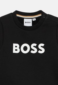 Crna pamučna majica s okruglim izrezom, s istaknutim bijelim logotipom "BOSS". Uključuje zlatno-bijelu prugastu detalj unutar ovratnika.