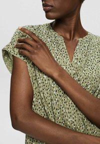 Žena v světle zeleném vzorovaném krátkém rukávu, která se dotýká opačného ramene, ukazuje výstřih a horní část trupu na neutrálním pozadí.