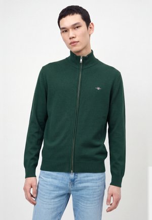 Un homme porte un pull vert foncé zippé à col montant et un jean bleu clair sur un fond blanc uni.