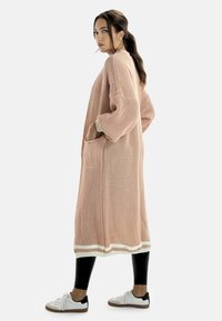 Rosa stickad kardigan med långa ärmar, sidofickor och vit kantning i nederkanten, kombinerad med svarta leggings och vita sneakers.