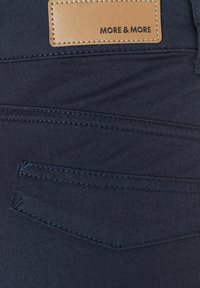 Tissu en denim bleu marine avec une étiquette en cuir beige portant l'inscription "MORE & MORE" et des coutures visibles le long du design de la poche arrière.