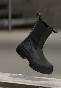 Schwarze Leder-Ankle-Boots mit elastischen Seiteneinsätzen, strukturiertem Obermaterial, dicker Gummisohle und Zuglaschen hinten, die über einer Oberfläche schwebend dargestellt sind.